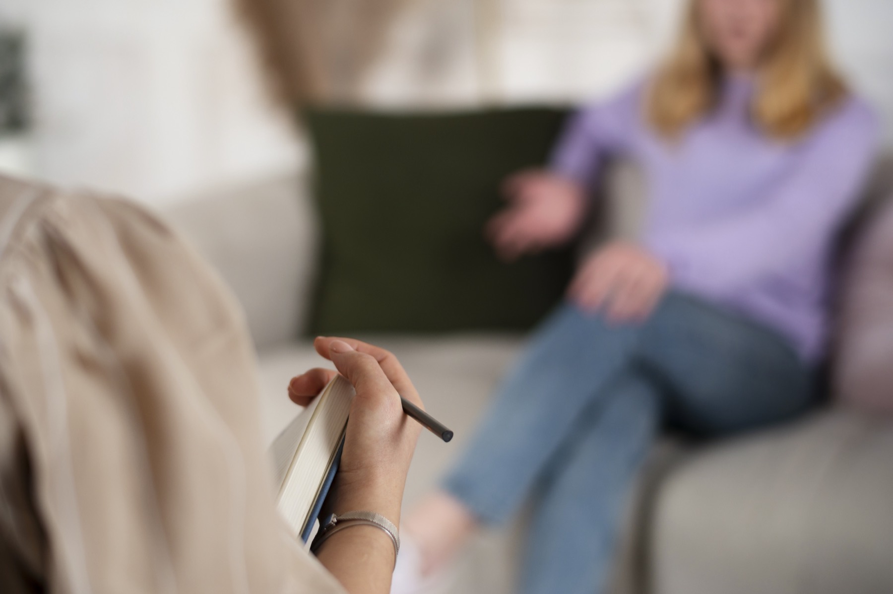 A therapist holding a notebook while a client speaks from the couch across from her.